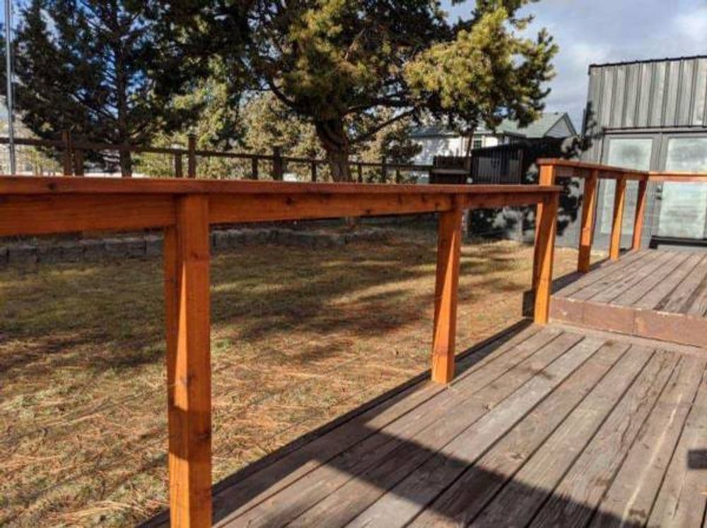 Wooden deck railing with a rustic finish, surrounded by trees and a clear blue sky.