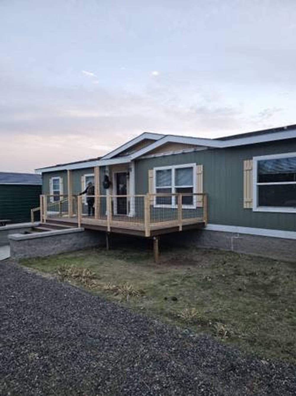 Modern green house with wooden deck and railing, surrounded by grass and gravel.