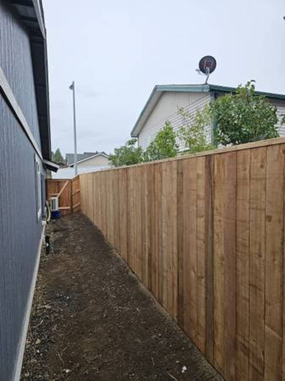 Wooden fence lined with plants next to a house under a cloudy sky.