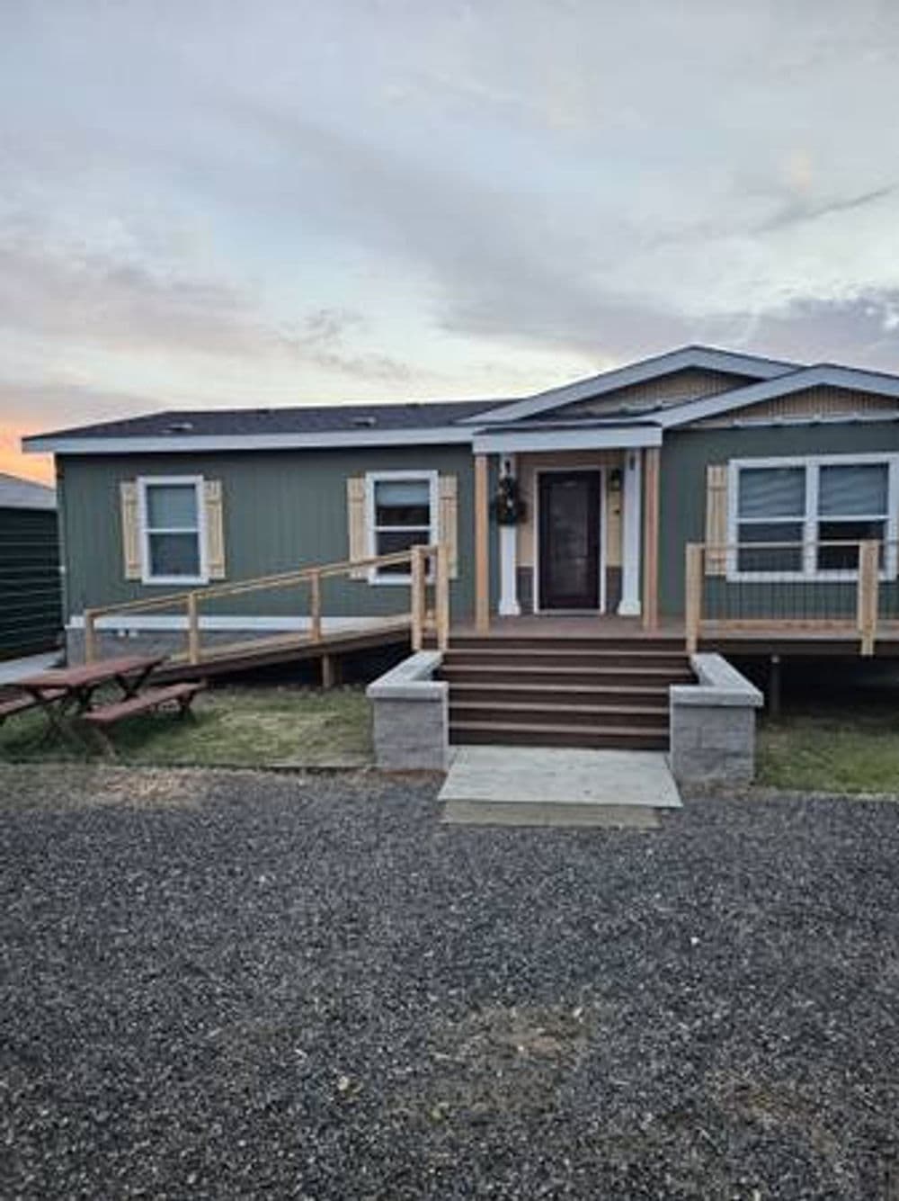 Modern green home with wooden deck, steps, and picnic area surrounded by gravel.