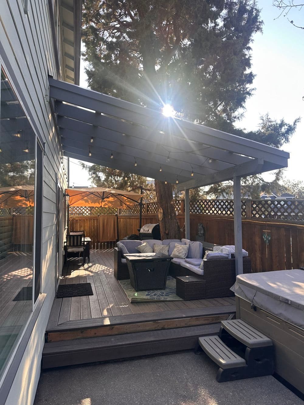 Cozy backyard patio with seating, string lights, and a hot tub under a shaded pergola.