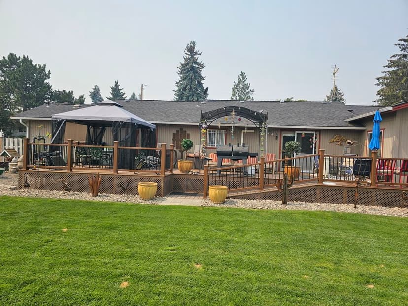 Backyard patio with deck, gazebo, and dining area, surrounded by greenery and sunny sky.