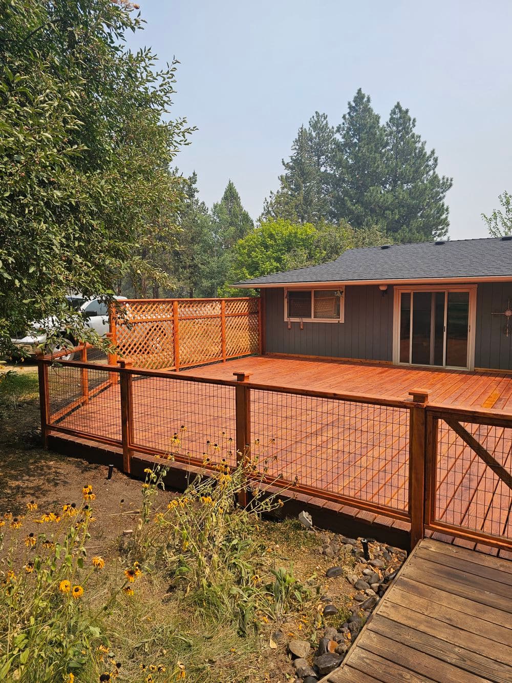 Wooden deck with lattice railing, surrounded by greenery and a house in the background.