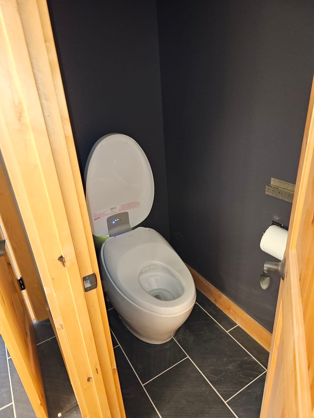 Modern toilet with open seat in a dimly lit bathroom, featuring wooden doors and gray tiles.