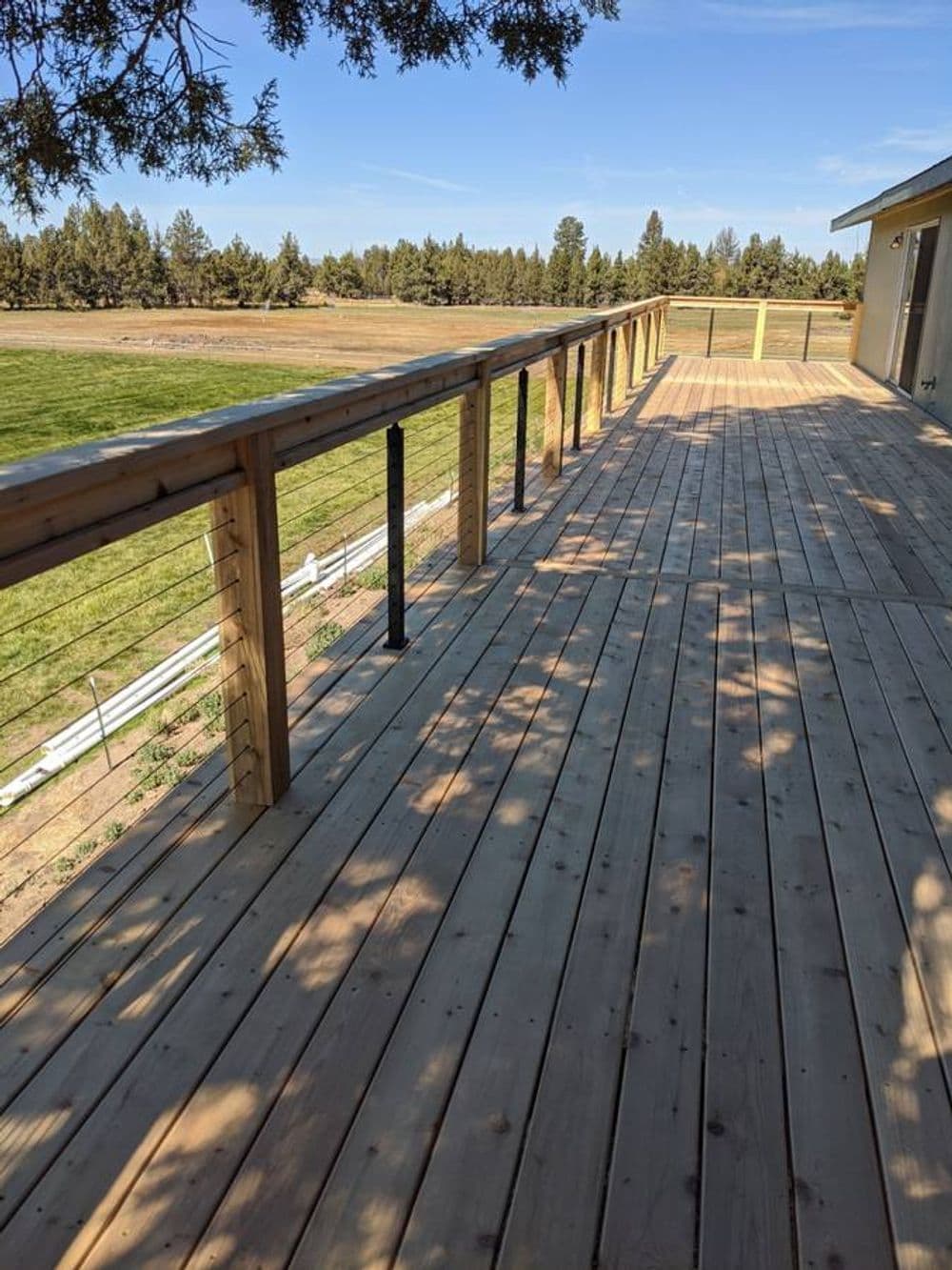 Spacious wooden deck with railing overlooking a grassy field and trees. Bright blue sky.
