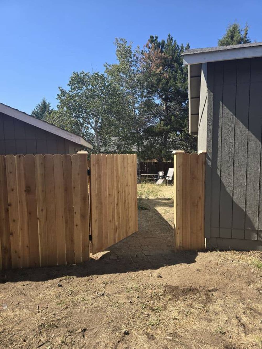 Open wooden gate between two gray houses leading to a grassy backyard area.