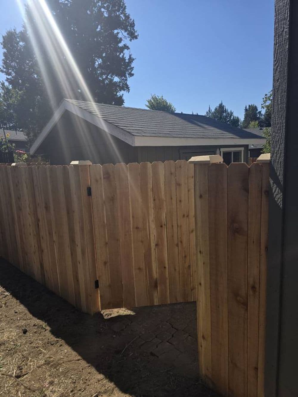 Wooden fence gate leading to a backyard with a house and trees under a clear blue sky.