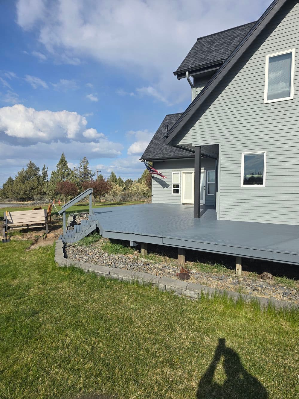 Modern gray house with a spacious deck and grassy yard under a blue sky with clouds.