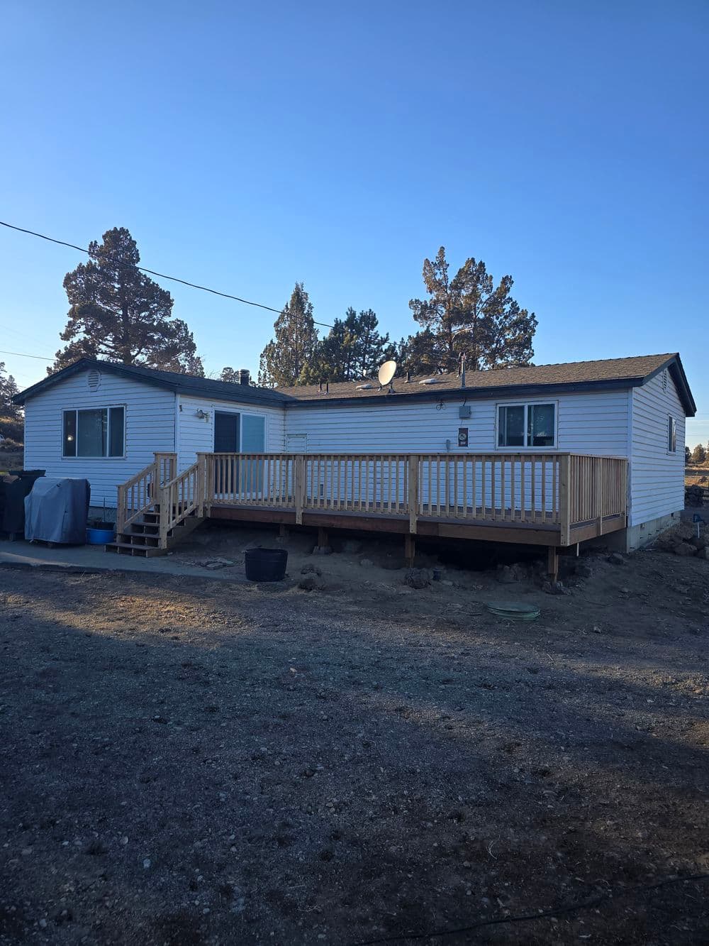 Single-story home with wooden deck, surrounded by trees and clear blue sky. Ideal for family living.