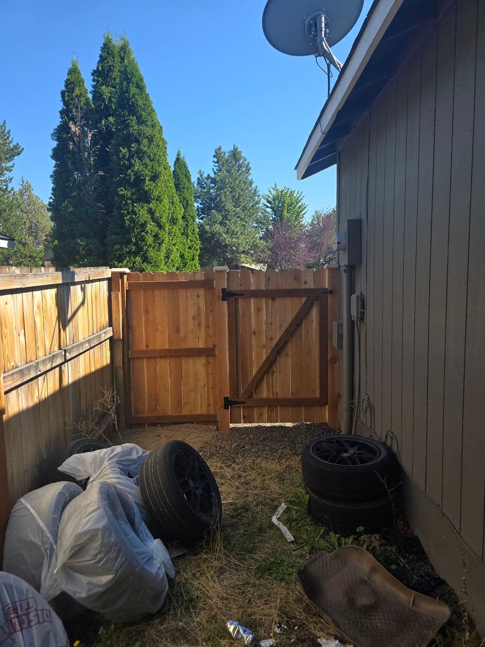 Side yard view with a wooden gate, tires, and a satellite dish under clear blue sky.