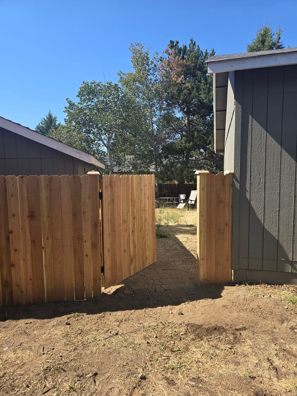 Open wooden gate between two grey houses leading to a clear backyard space.
