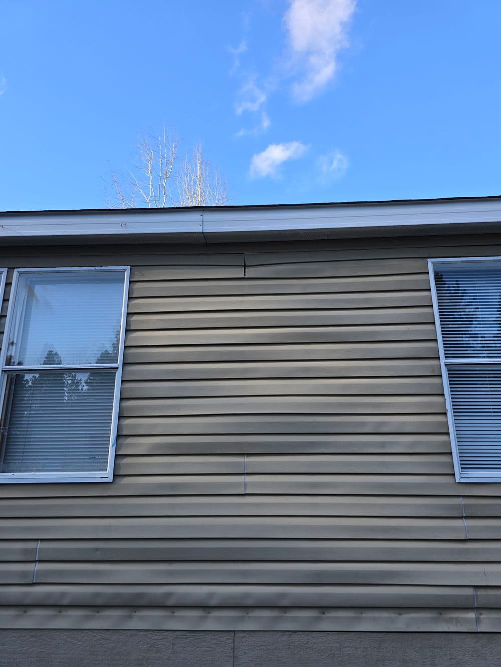 Side exterior of a house with vinyl siding and clear blue sky. Visible window and slight damage.