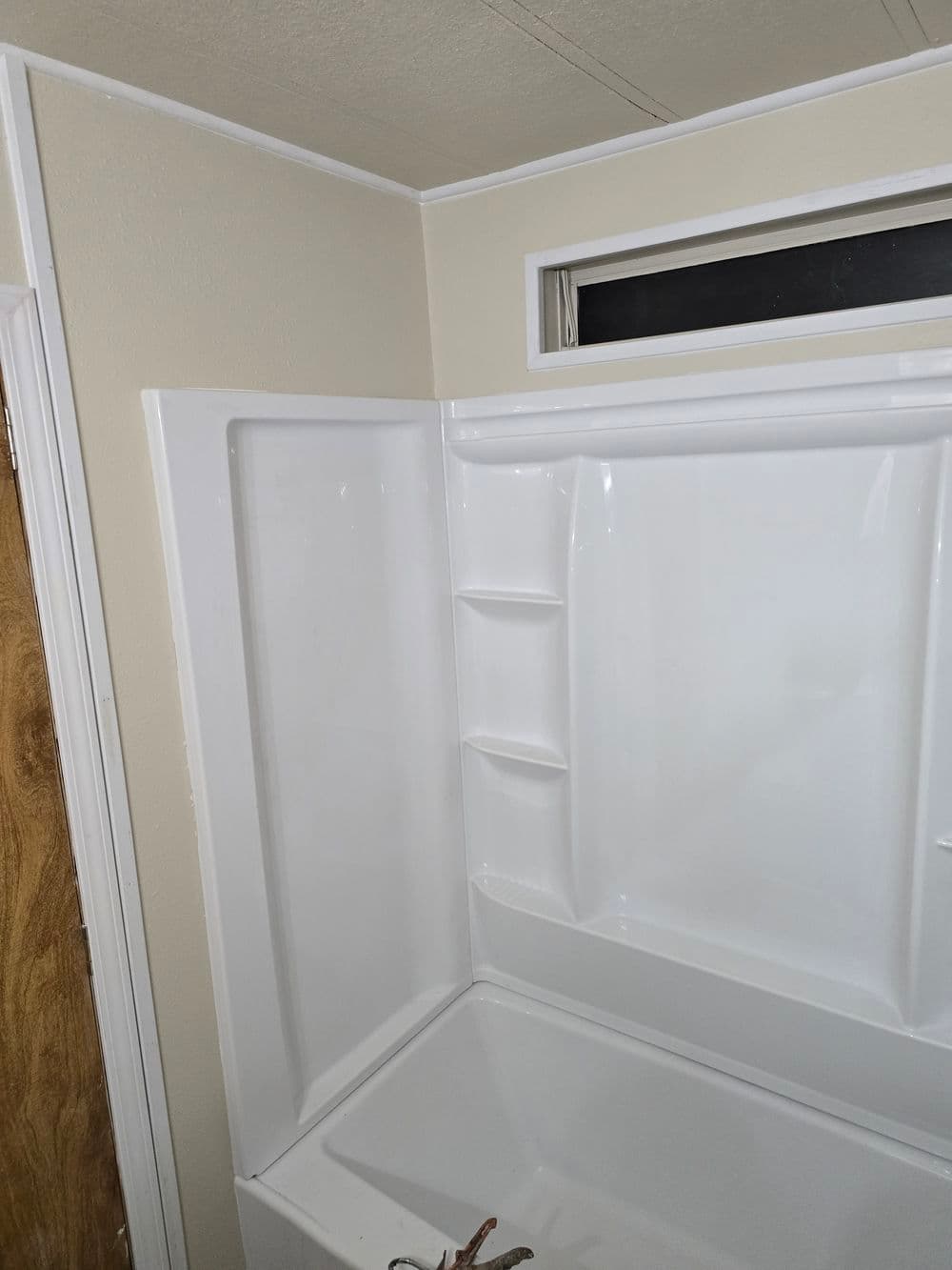 Newly installed white acrylic bathtub and shower enclosure with shelving and window.