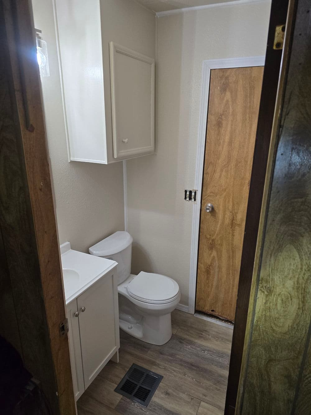 Small bathroom with white cabinet, toilet, and wooden door, featuring light wood flooring.