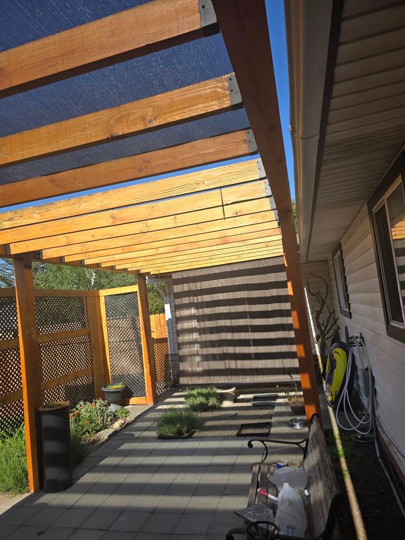 Wooden pergola with shaded area over pavers, adjacent to a house and garden features.