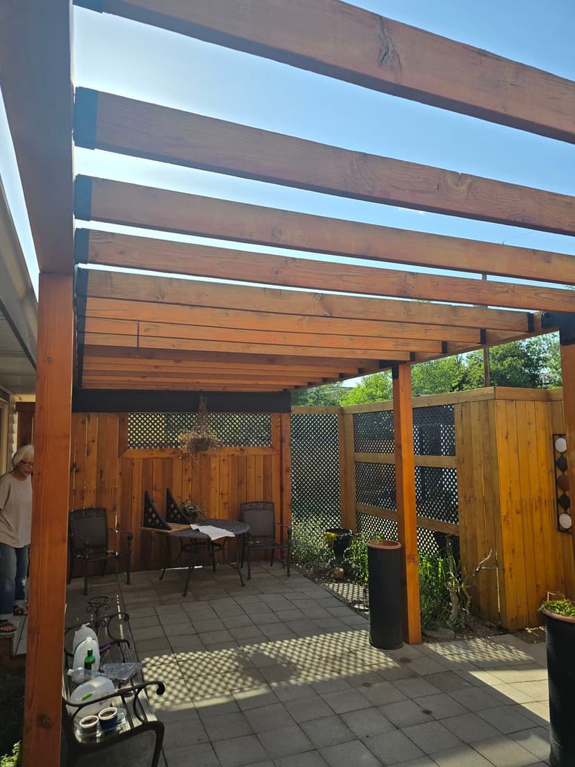 Wooden pergola in a patio setting with chairs and a table under blue sky.