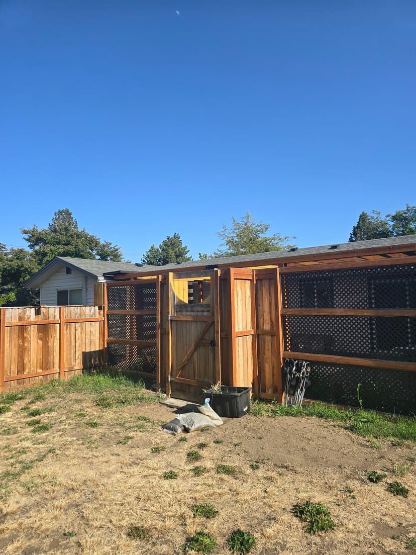 Wooden fenced yard with a gate, surrounded by greenery and a clear blue sky.